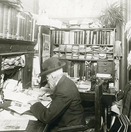 \"A black and white image of a man working at a desk filled with papers and books, c. 1910. "