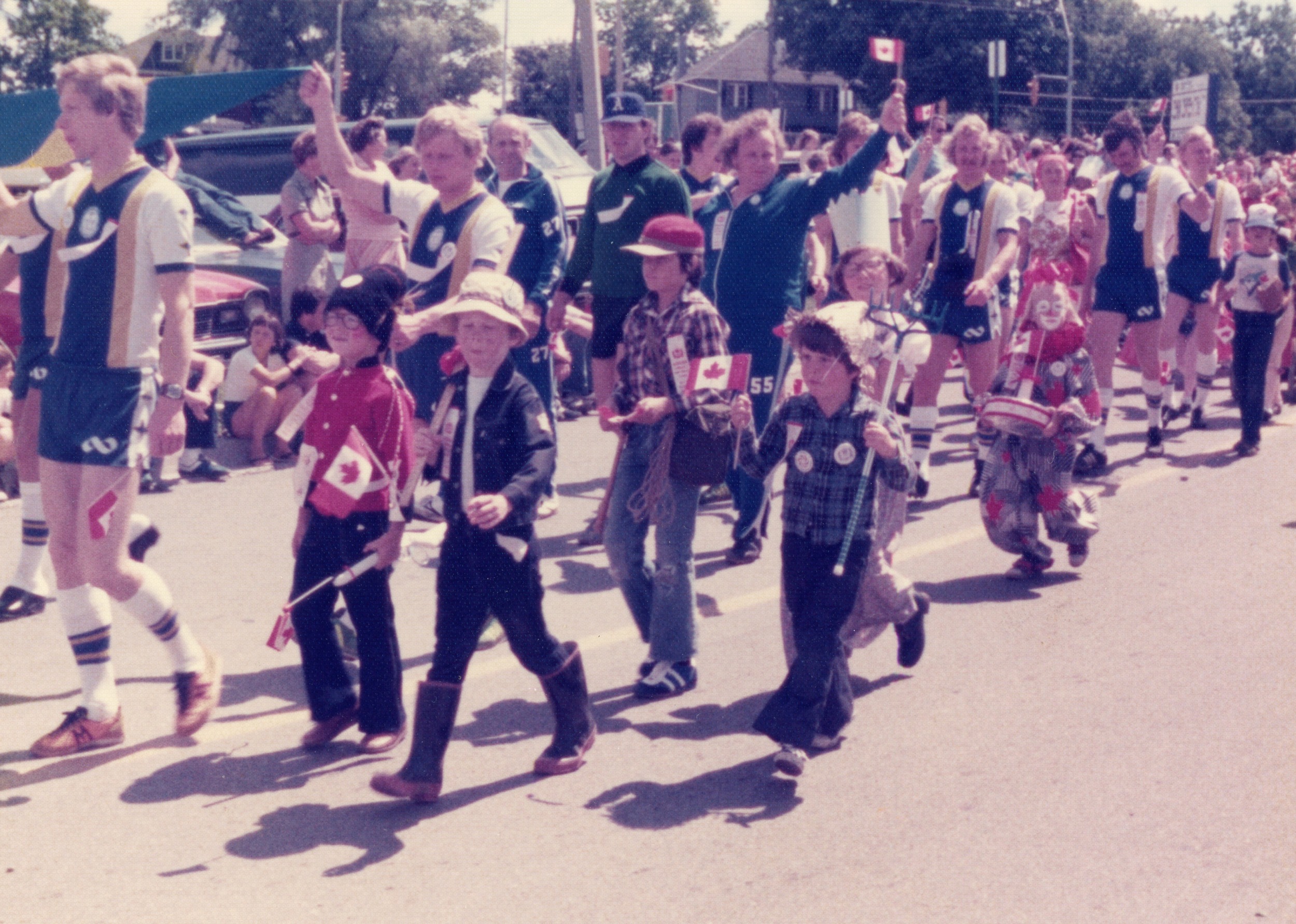 "A colour photo of kids of various ages walking down the road participating in a Canada Day parade, 1977"