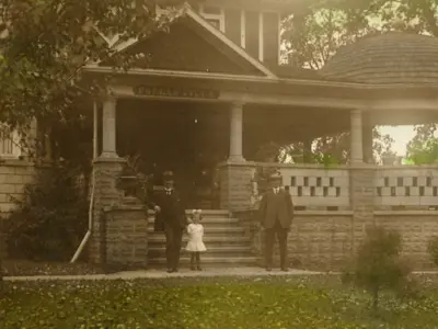Very old picture of a home, surrounded by trees, with three people standing in front