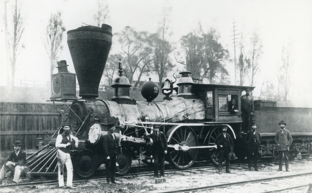 A black and white photo of an old steam-engine with 9 men standing in and around it.
