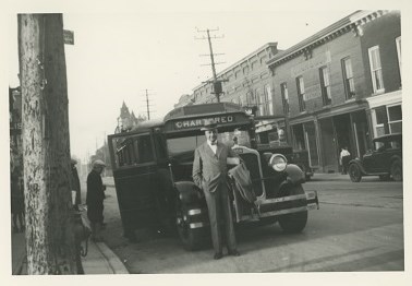Black and white photograph a 1920s street scene showing a school bus parked on the site of the road with a man standing in front of it.